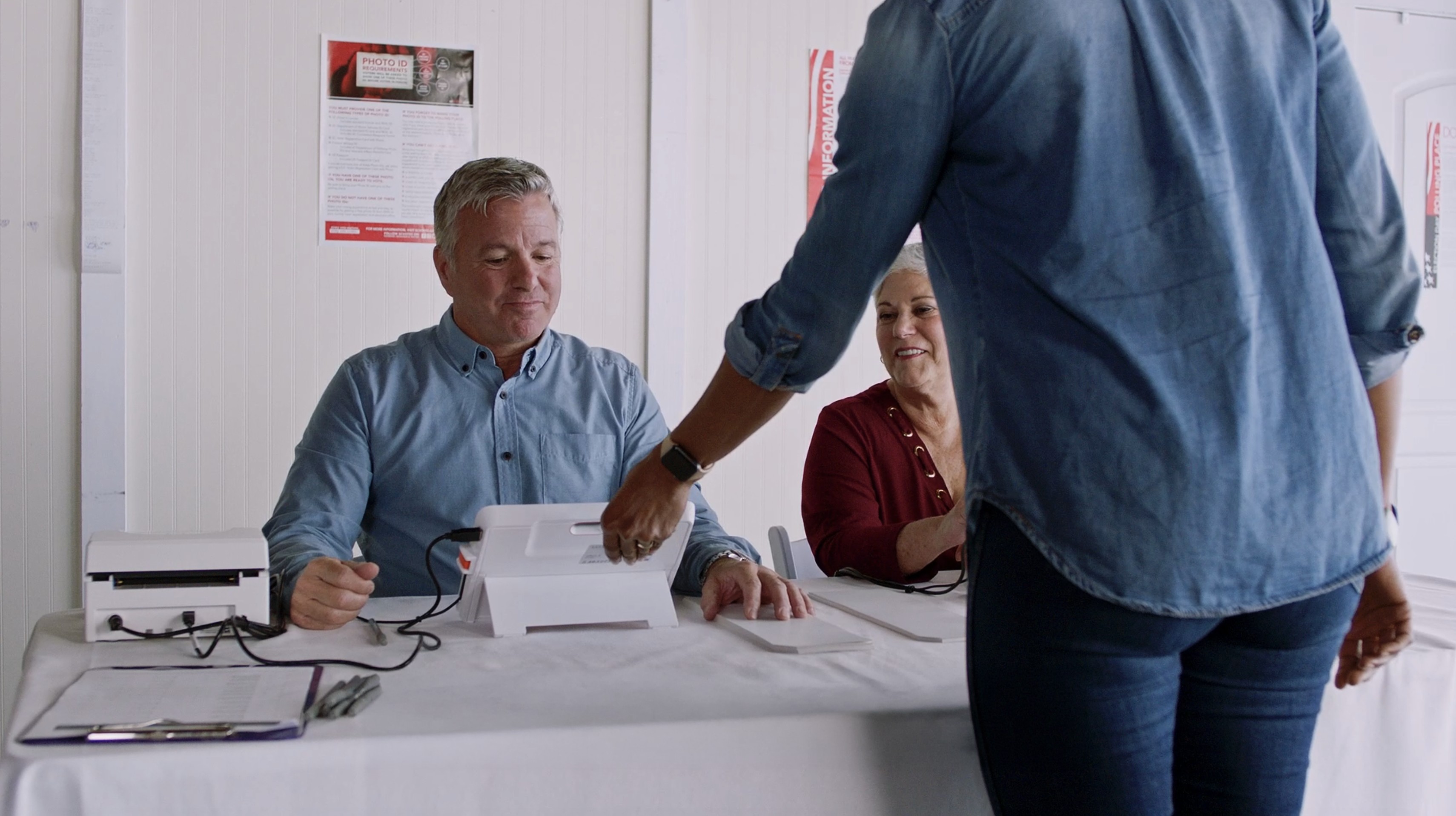 A poll worker assists a voter by handing them a voting tablet at a polling place while another poll worker looks on.